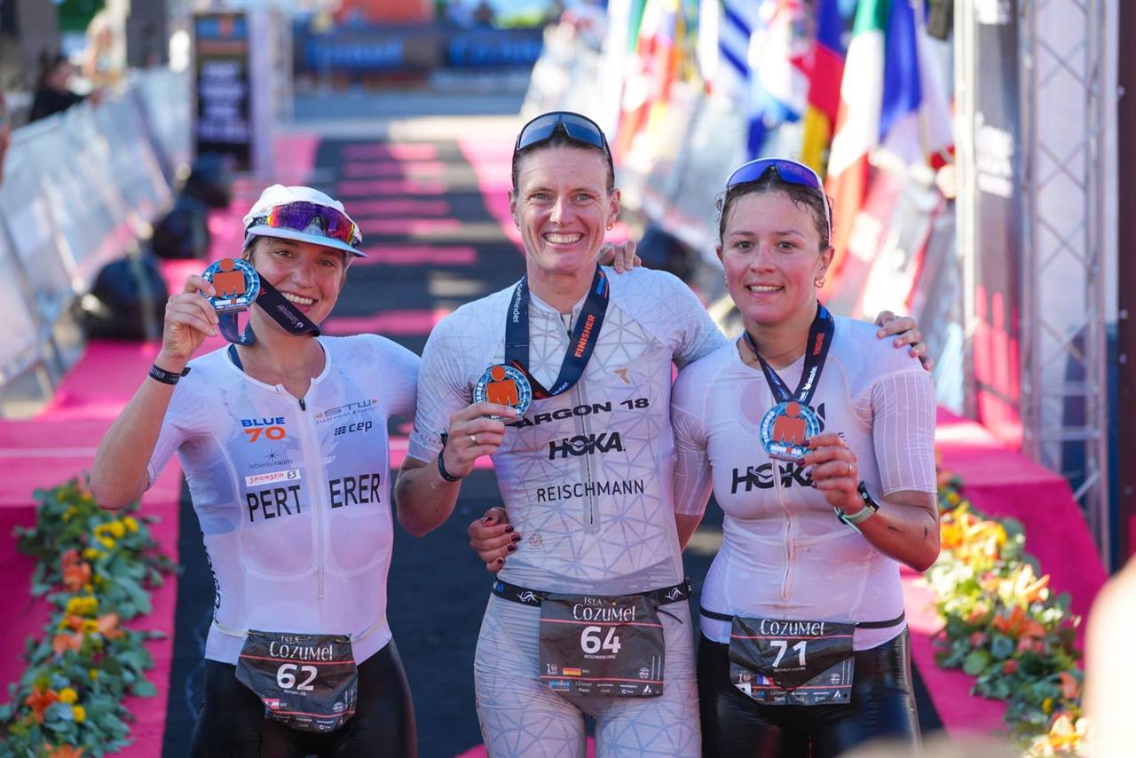 Three women triathletes smiling with their finisher medals at the IRONMAN Cozumel finish line